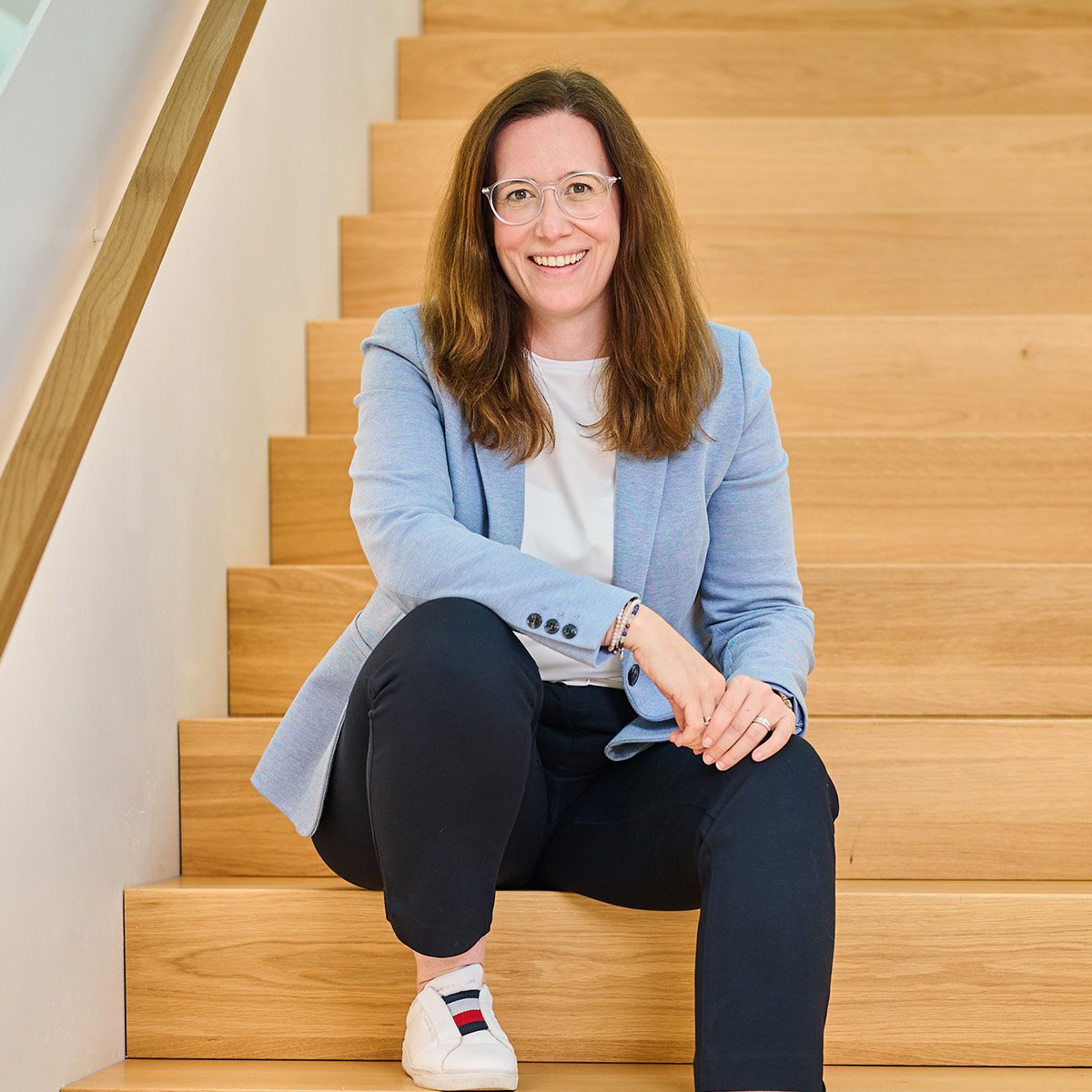 Maike Abel: Die Fotografie zeigt eine lächelnde Frau mit schulterlangem braunem Haar und heller Haut, die auf einer breiten Holztreppe sitzt. Sie trägt eine helle Brille, ein hellblaues Sakko über einem weißen Shirt, eine dunkle Hose und weiße Sneakers mit rot-blauem Streifen. Ihr rechter Arm ruht locker auf ihrem rechten Knie, während sie mit der linken Hand das andere Bein berührt. Der Hintergrund zeigt die helle Holzstruktur der modernen Treppe, deren Geländer links im Bild verläuft. Die Szene wirkt freundlich, professionell und offen.