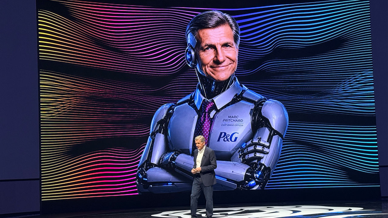 Speaker on stage in front of a large screen showing a futuristic robot with visible text “P&G” and “Marc Pritchard Chief Brand Officer”.