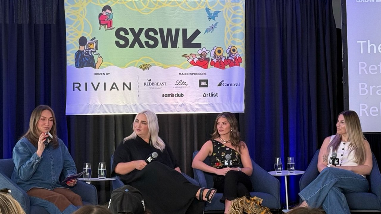 Four people seated on stage at a SXSW panel in front of a banner with the SXSW logo; one person speaks into a microphone while the others listen.