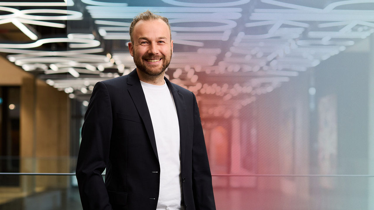 Portrait of Mario Wimmer in front of a blurred indoor background. He is smiling and wearing a dark blazer over a white shirt.