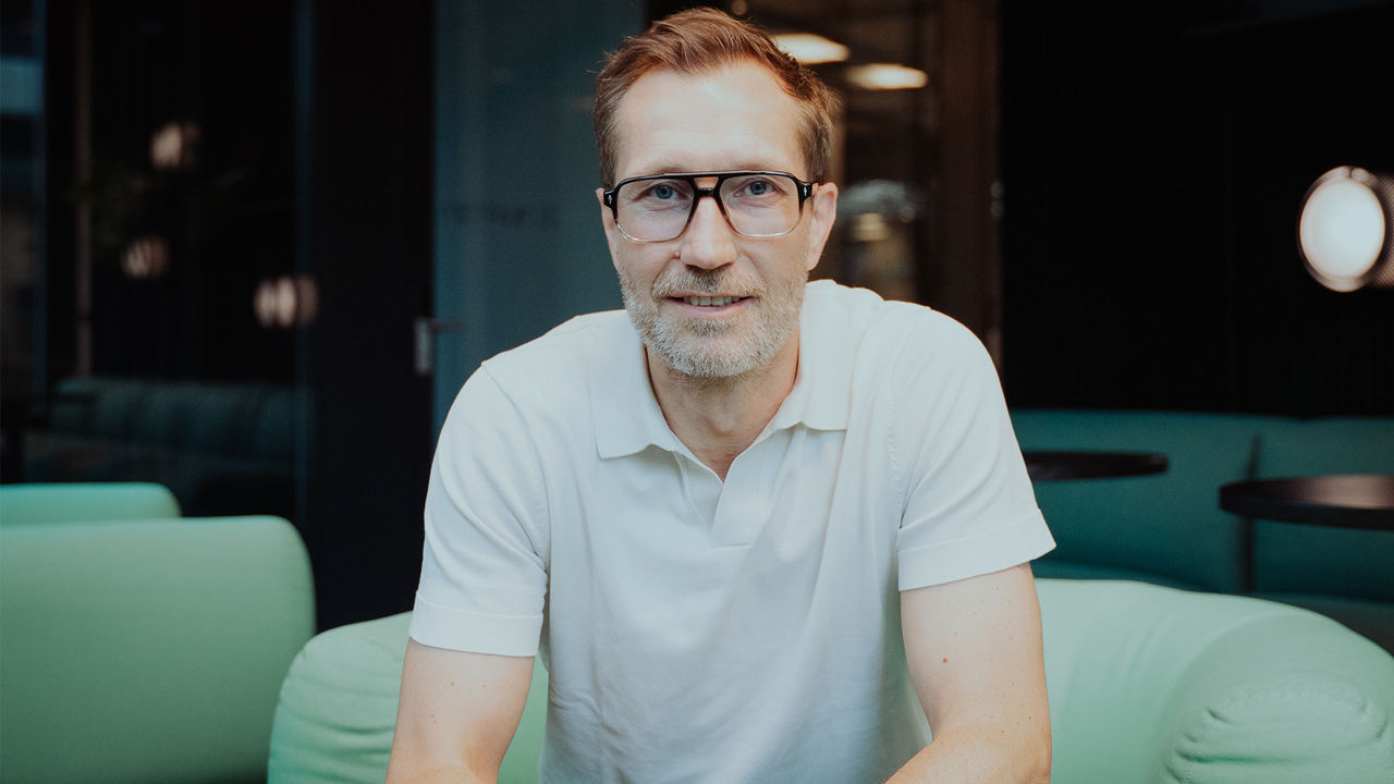 Portrait of a man wearing glasses and a white polo shirt, seated indoors and looking toward the camera. The background shows a modern office or lounge setting with soft lighting and blurred furnishings.