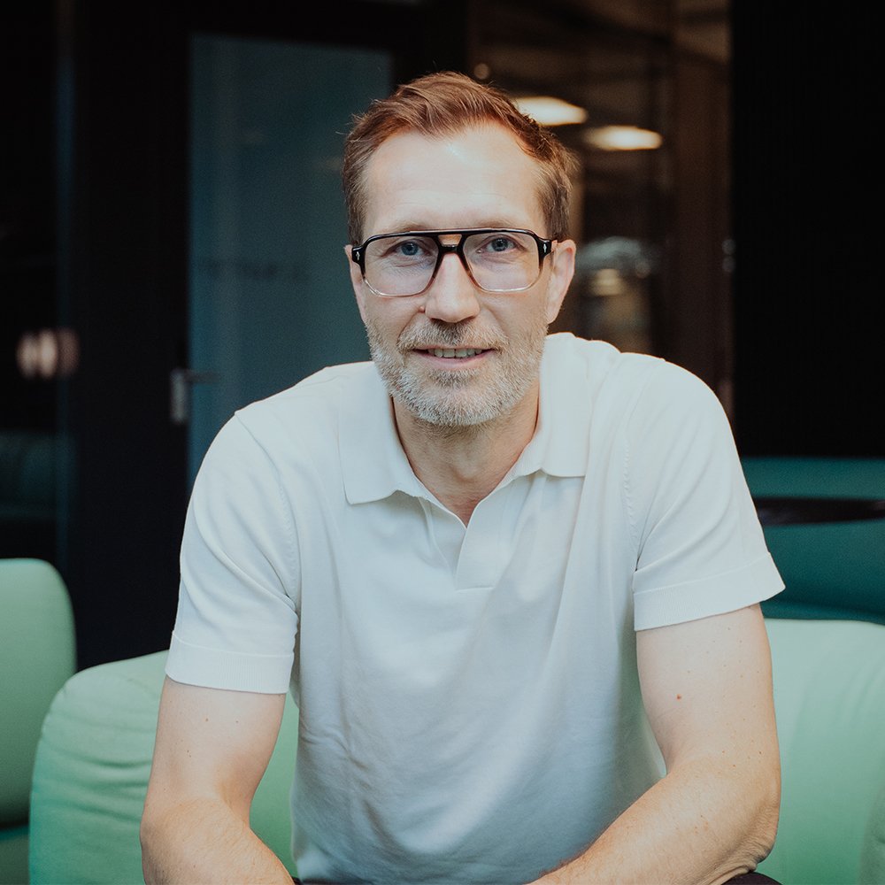 Portrait of a man (Markus Noder) wearing glasses and a white polo shirt, seated indoors and looking toward the camera. The background shows a modern office or lounge setting with soft lighting and blurred furnishings.