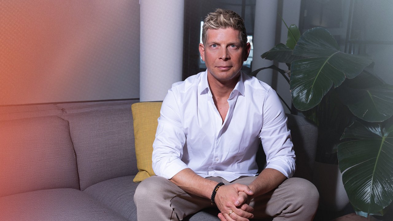 Portrait of Max Schöngen in an indoor setting with a plant in the background. He is wearing a white shirt and looking directly at the camera.