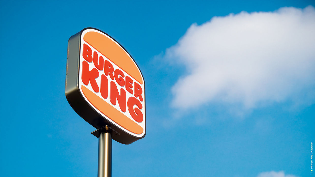 Close-up of a Burger King sign with the iconic red and orange retro-style logo mounted on a tall pole, photographed in daylight against a clear blue sky with a single white cloud. The image represents the visual branding of the global fast-food chain Burger King.