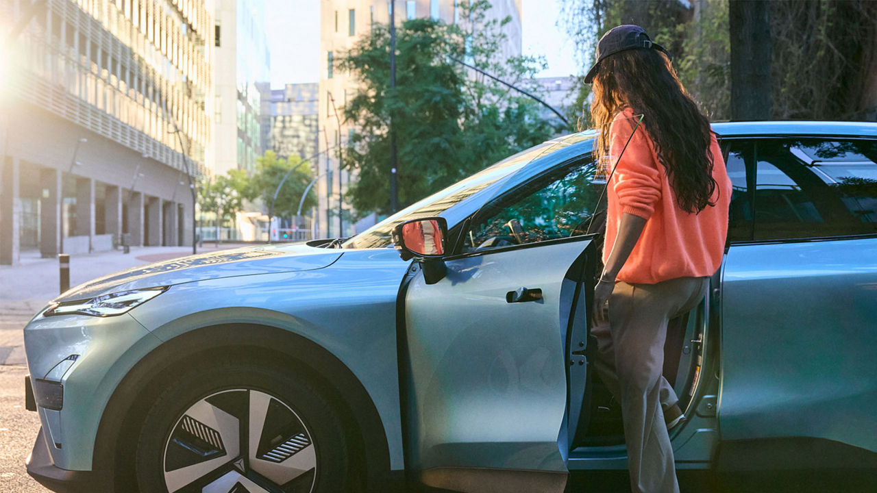 A person with long dark hair wearing an orange sweater and a cap is getting into a blue Changan car on a sunny city street. The driver’s door is open, with modern buildings and trees visible in the background.