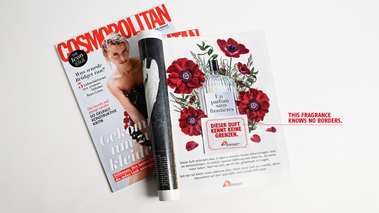 An open copy of *Cosmopolitan* magazine lies on a light-colored surface. The right-hand page features an advertisement showing a perfume bottle, red flowers, and the slogan “This fragrance knows no bounds.” At the bottom of the page is additional information about the organization Doctors Without Borders. The left-hand page shows the magazine’s cover, which features a person.