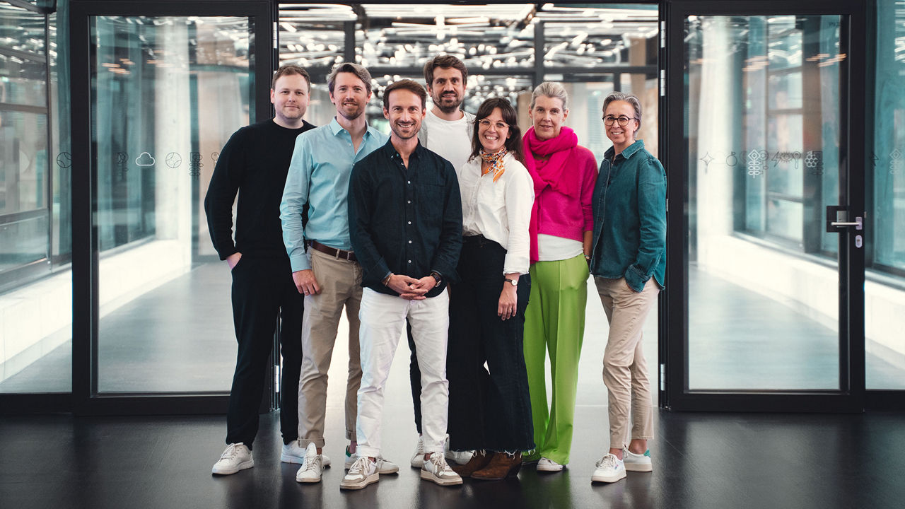 Seven people are standing side by side, smiling, in a modern office hallway with glass walls and a dark floor. They are dressed in smart casual attire, with some wearing colorful pieces like green pants or a bright pink sweater. Light structures are reflected in the ceiling behind them.
