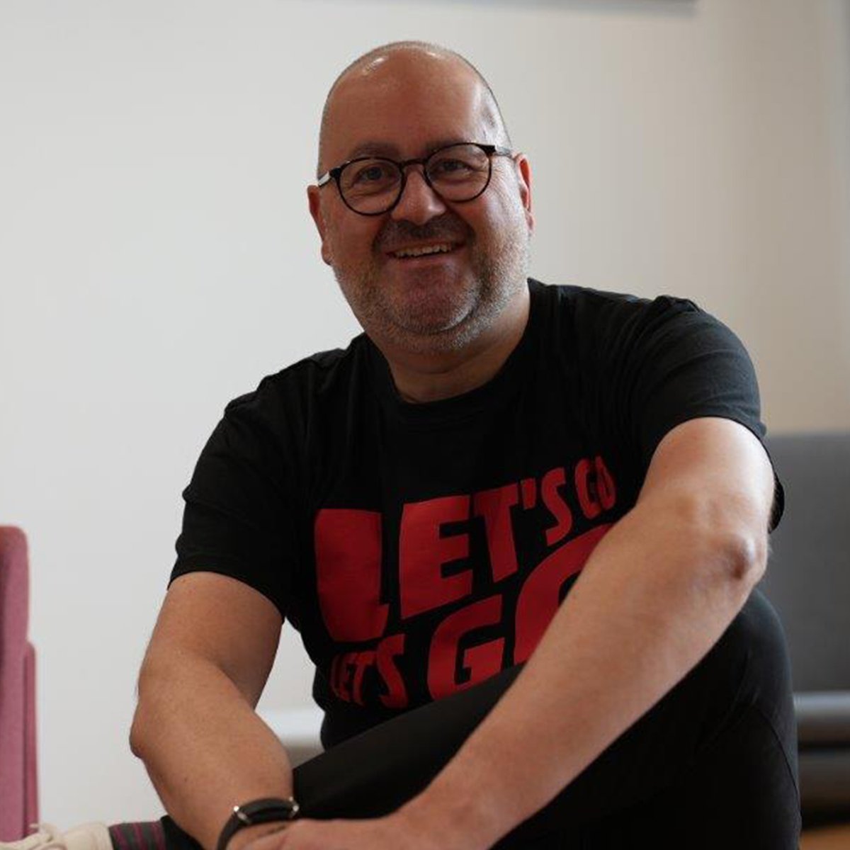 Michael Schuld: Ein Mann mittleren Alters mit Glatze, Brille und kurzem Bart sitzt locker auf einem rosa Stuhl. Er trägt ein schwarzes T-Shirt mit rotem Schriftzug „LET’S GO“, eine schwarze Hose und weiße Sneaker mit MediaMarkt-Logo. Im Hintergrund sind unscharf die Logos von MediaMarkt und Saturn an der Wand zu erkennen. Die Atmosphäre wirkt entspannt und freundlich.