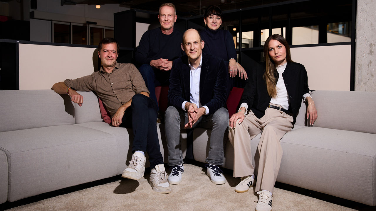 Fünf erwachsene Personen sitzen und stehen gemeinsam in einem modernen Innenraum mit Sofas und Glaswänden. Sie blicken in die Kamera und tragen überwiegend dunkle, legere Kleidung.