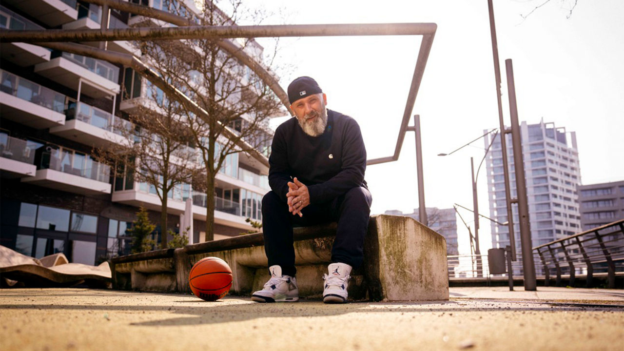 Person mit Kappe sitzt auf einer Betonbank im Außenbereich neben einem Basketball, im Hintergrund moderne Wohngebäude.