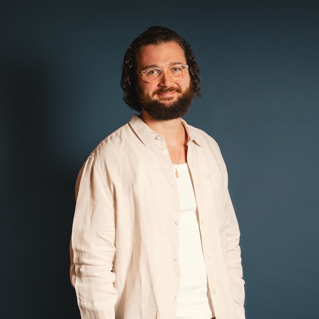 Porträt eines Mannes mit lockigen Haaren, Bart und Brille vor blauem Studiohintergrund. Er trägt ein helles Hemd und lächelt freundlich in die Kamera.