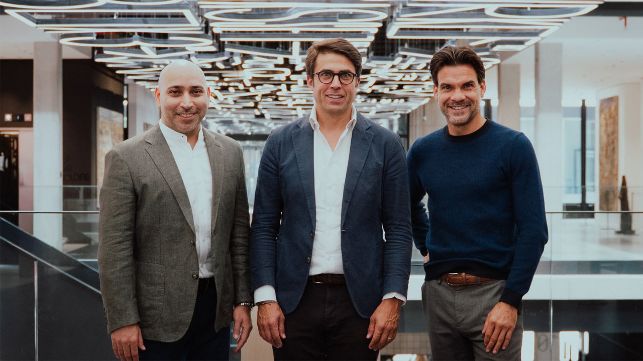 Three people in business-casual attire standing in a modern office building with distinctive ceiling lighting.