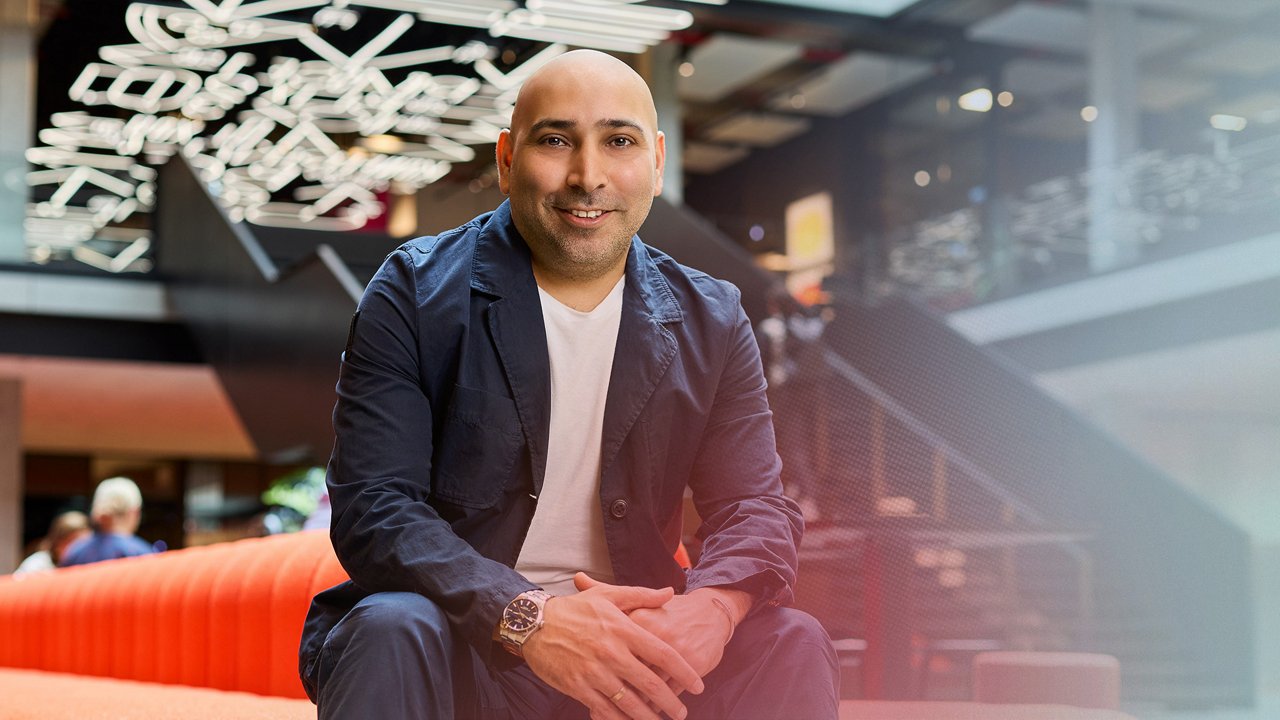 Porträtaufnahme von Hadi Lotfi in dunkler Jacke und hellem T-Shirt. Die Person sitzt in einem modern gestalteten Innenraum mit unscharfen Lichtinstallationen im Hintergrund.