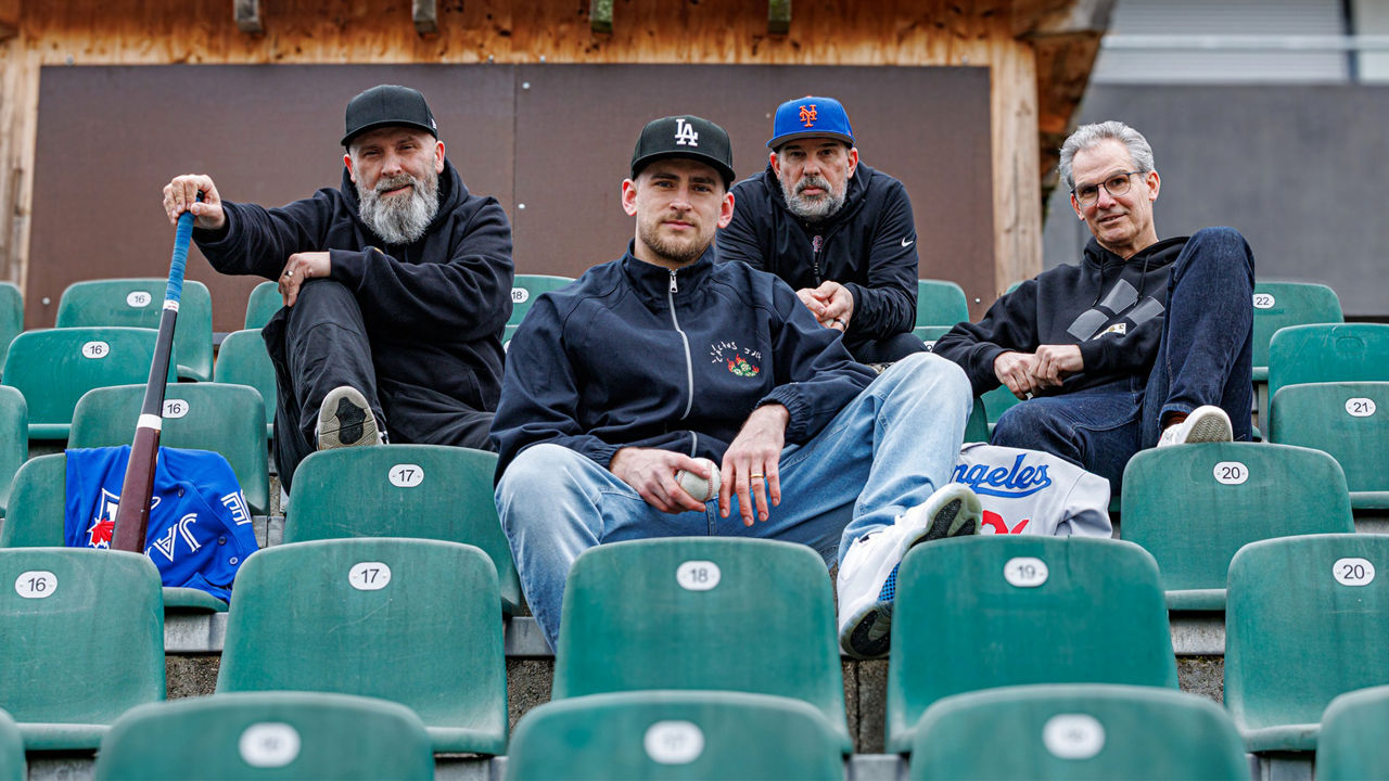 Vier Personen sitzen in den Zuschauerreihen eines Baseballstadions; eine Person hält einen Baseball, neben ihnen liegt ein Baseballschläger.