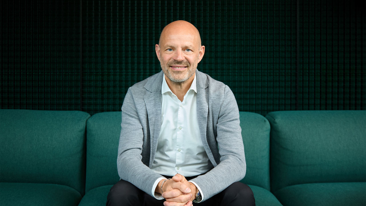 Porträt eines sitzenden Mannes im hellen Sakko auf einem grünen Sofa vor dunklem Hintergrund.