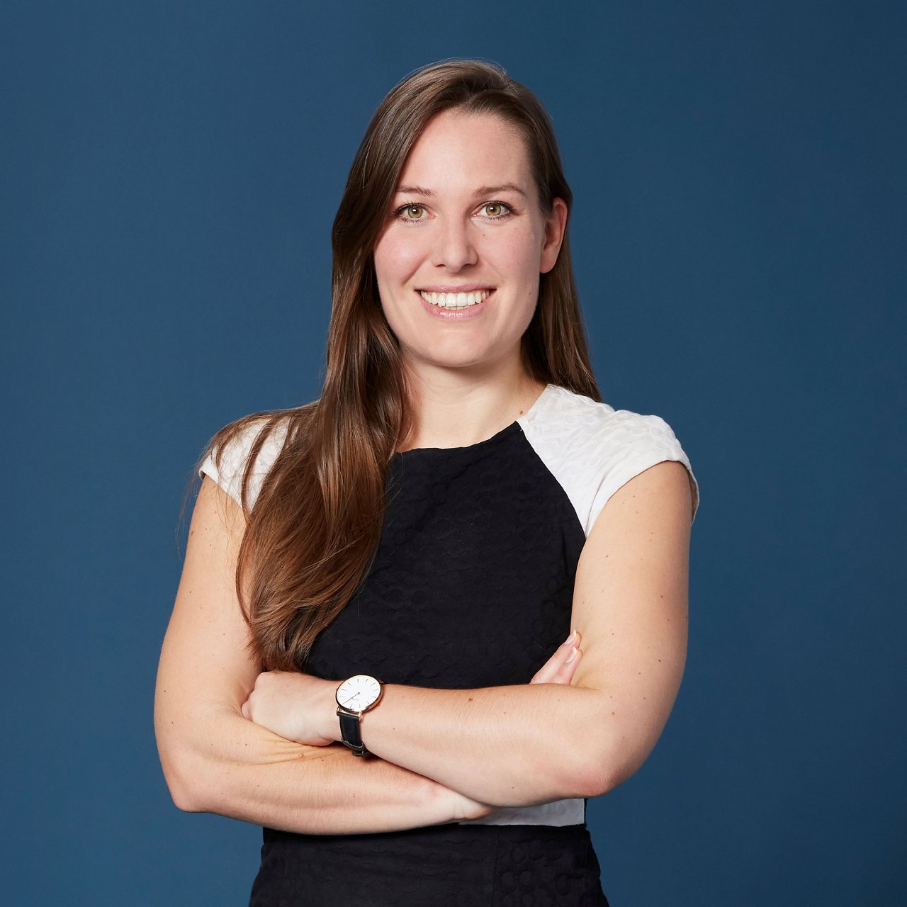 Katrin Krall steht vor einem einfarbig blauen Hintergrund, lächelt in die Kamera und hat die Arme vor der Brust verschränkt. Sie trägt ein ärmelloses Kleid in Schwarz und Weiß sowie eine Armbanduhr.
