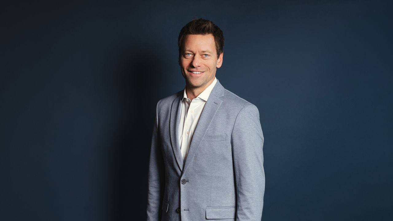 Philipp Brunner stands in front of a dark blue background. He is wearing a light blue blazer over a white shirt and looks at the camera with a slight smile. The image is clearly lit, with the subject centered against a plain backdrop.