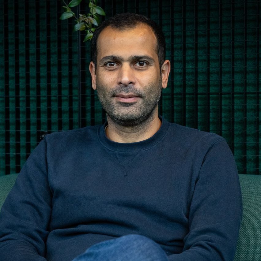 Hatem Fahik: a man with short dark hair and a trimmed beard is sitting on a green couch in front of a dark textured wall. He is wearing a navy blue sweatshirt and jeans, looking at the camera with a neutral expression.