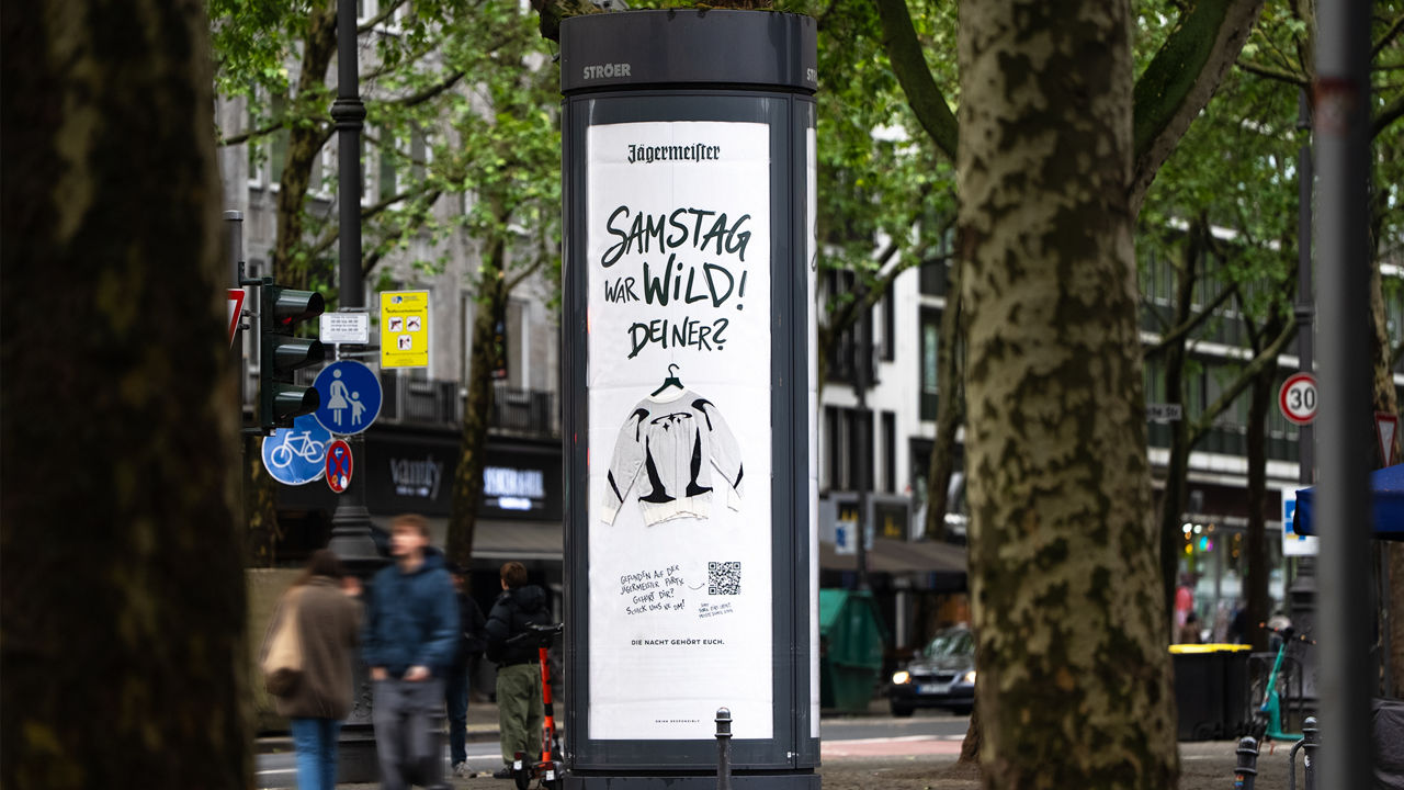 Digitale Litfaßsäule in einer begrünten Innenstadtstraße mit Jägermeister-Werbung und dem Schriftzug „Samstag war wild! Deiner?“ neben einer aufgehängten Jacke.