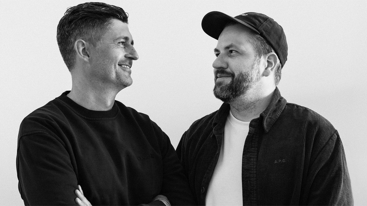 The image shows Jan-Philipp Jahn and Till Diestel: The black-and-white portrait depicts two men looking at each other in a friendly manner, standing turned toward one another; the man on the left is wearing a dark sweatshirt with his arms crossed, while the man on the right is wearing a corduroy jacket, a T-shirt, and a baseball cap