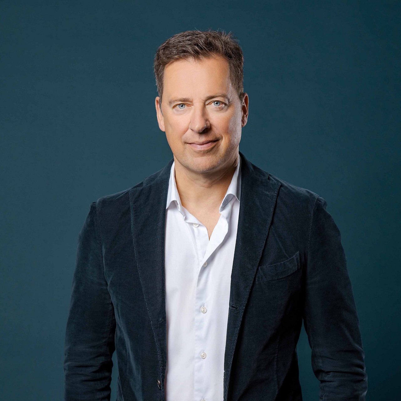 Matthias Brüll, a man with short brown hair and fair skin, stands in front of a dark blue background. He is wearing a white shirt and a dark blue corduroy jacket. He looks directly at the camera with a slight smile