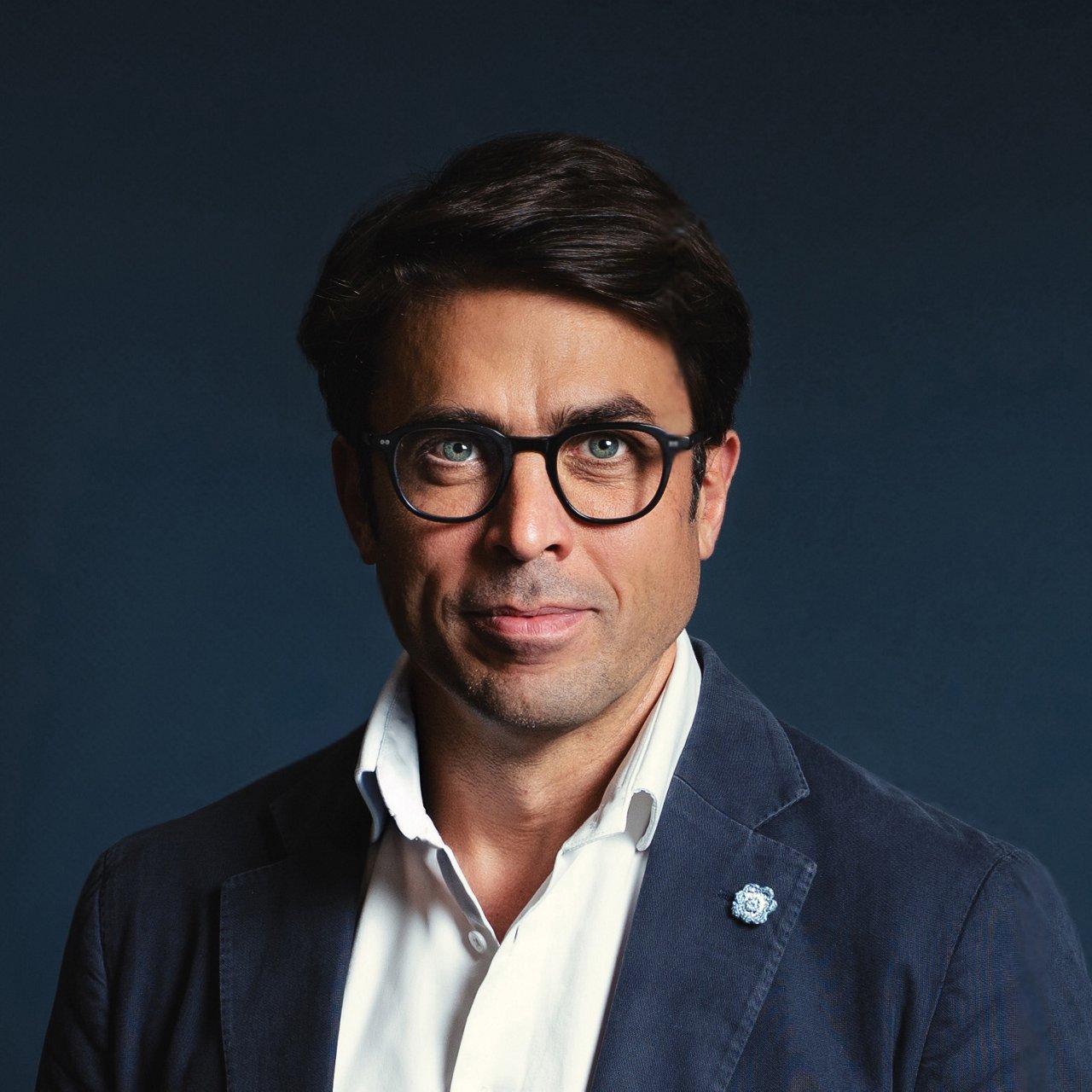 Christian Schmitz, a man with short dark hair, fair skin and dark-rimmed glasses, is looking at the camera with a friendly expression. He is wearing a white shirt and a dark blue blazer with a lapel pin. The background is solid dark blue.