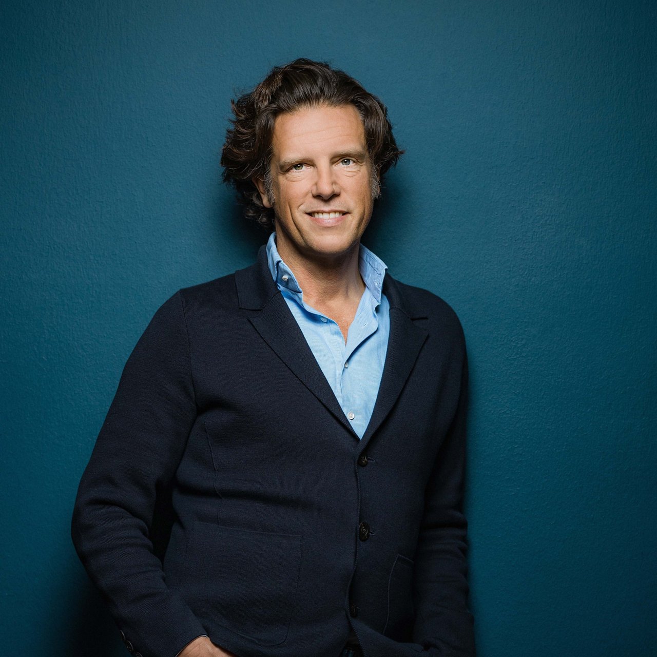 Florian Haller, a man with medium-length brown hair and fair skin, is smiling at the camera. He is wearing a light blue shirt and a dark blue jacket, standing casually with his hands in his pockets against a teal-colored wall.