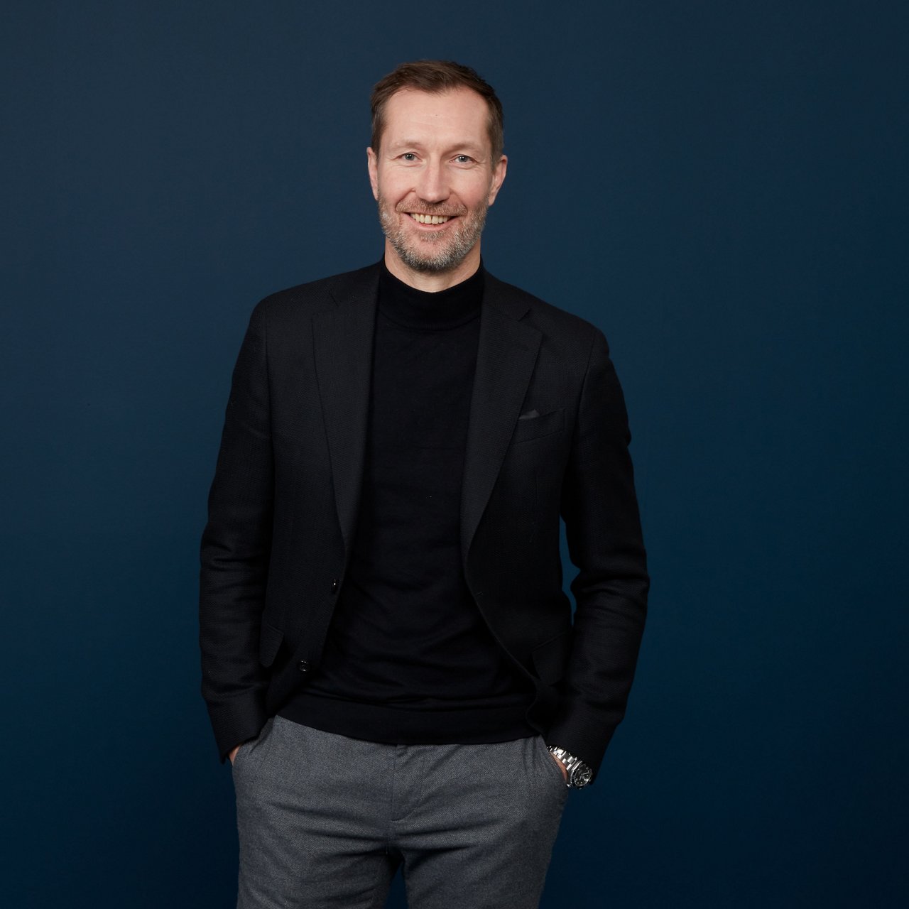 Markus Noder, a man with short brown hair, fair skin, and a light beard, stands smiling in front of a dark blue wall. He is wearing a black blazer with a black turtleneck and gray trousers, with his hands in his pockets.