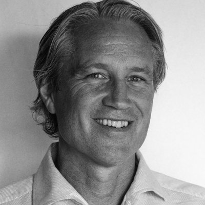 Schwarz-weiß-Porträt eines lächelnden Mannes mit leicht gewelltem, nach hinten gekämmtem Haar. Er trägt ein helles Hemd und blickt direkt in die Kamera.