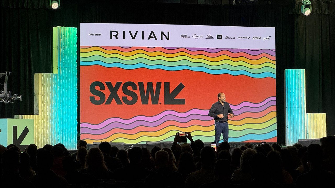 A speaker stands on a SXSW stage in front of a large screen with the SXSW logo while addressing an audience.