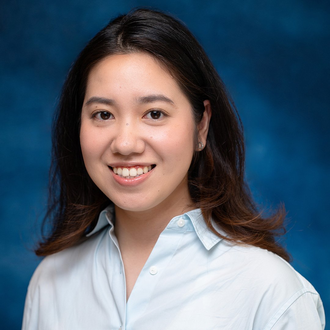 Porträt einer Frau mit schulterlangen dunklen Haaren und hellblauer Bluse vor blauem Hintergrund. Sie lächelt freundlich und blickt direkt in die Kamera.