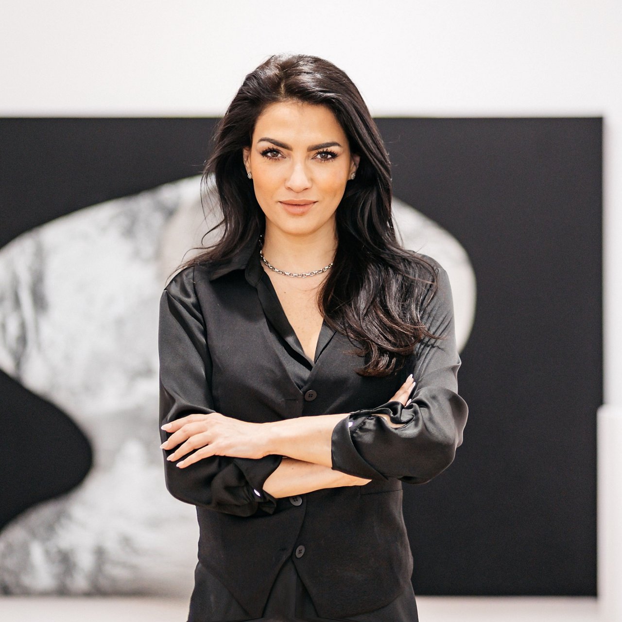 Ruth Santana, a woman with long dark brown hair and fair skin, stands with her arms crossed in front of a modern black-and-white artwork. She is wearing a black blouse and a silver necklace. The background features a light gallery wall with minimalist art.