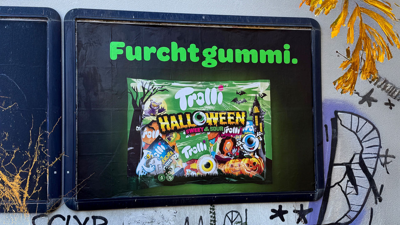 Plakatwerbung von Trolli mit dem Schriftzug „Furchtgummi.“ in leuchtend grüner Schrift auf schwarzem Hintergrund. Darunter ist eine Packung „Trolli Halloween Sweet & Sour“ abgebildet, geschmückt mit Halloween-Motiven wie Kürbissen, Fledermäusen, Gespenstern und einem gruseligen Auge. Das Plakat kombiniert Wortwitz und Halloween-Optik zu einer humorvollen Süßigkeitenkampagne