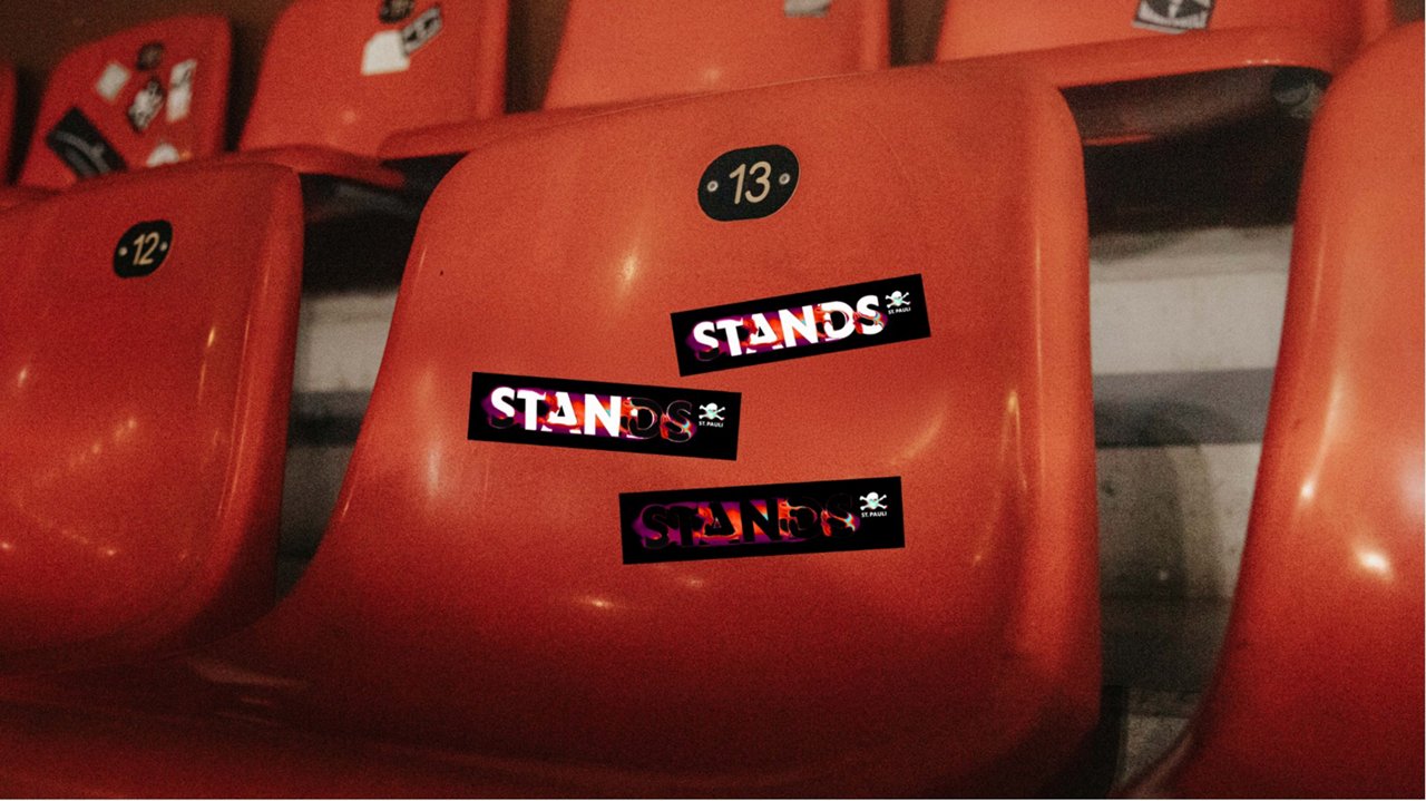 Rote Stadionschalen mit den Sitznummern 12 und 13. Auf dem Sitz Nr. 13 kleben drei schwarze Sticker mit dem leuchtenden Schriftzug „STANDS“ und dem St. Pauli-Totenkopf-Logo.