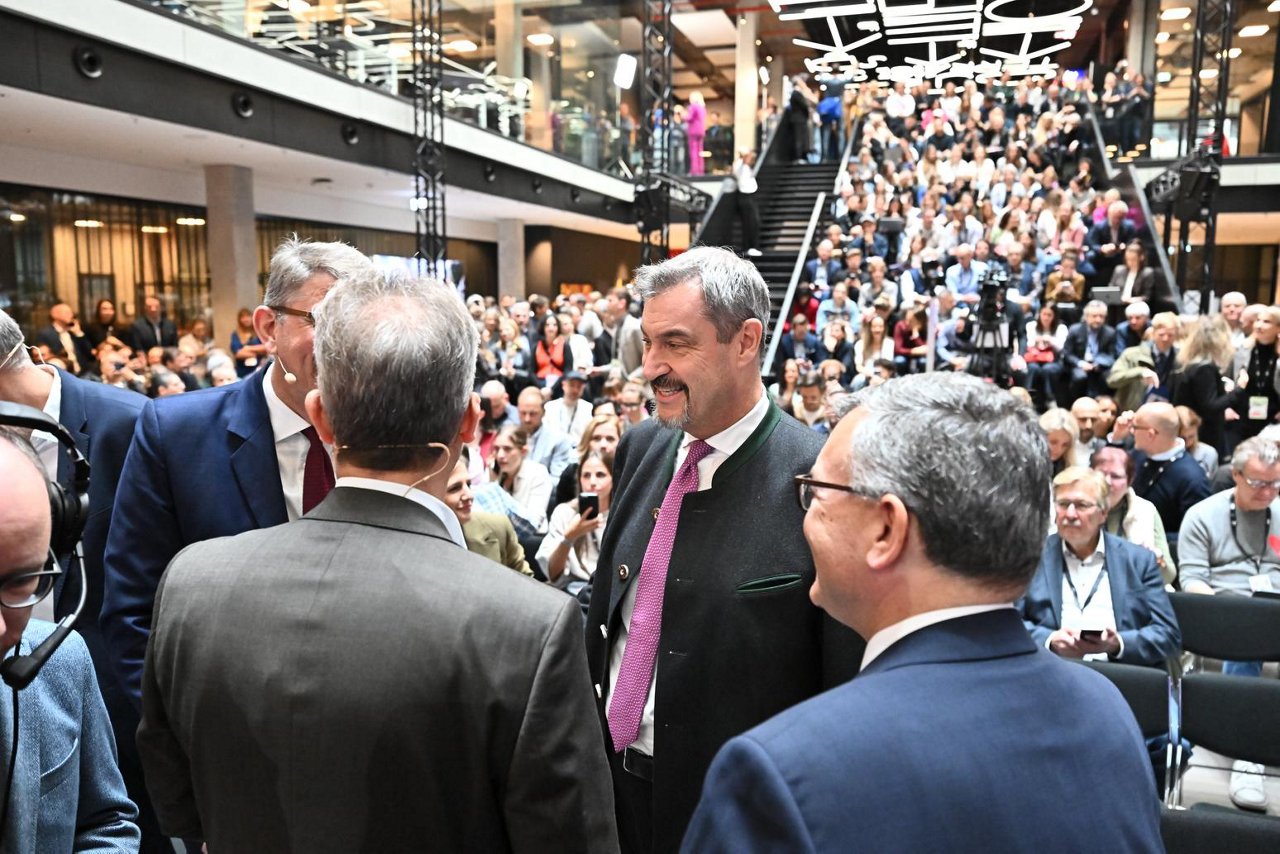 Das Bild zeigt eine lebhafte Konferenzszene im House of Communication in München. Im Vordergrund stehen mehrere Personen in angeregtem Gespräch, während im Hintergrund zahlreiche Gäste auf einer Treppe und Sitzplätzen Platz genommen haben. Moderne Architektur und Beleuchtung prägen die Atmosphäre.
