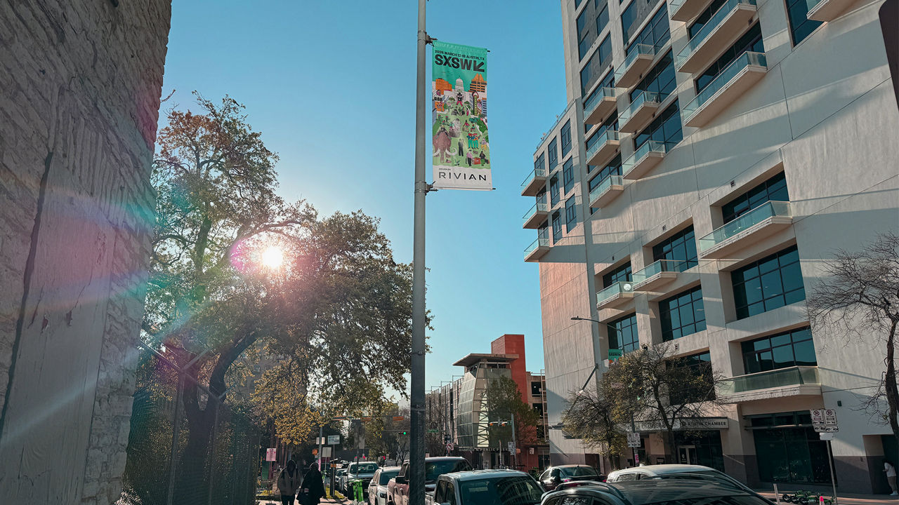 Sunny city street with parked cars, trees, and modern buildings. A banner for the SXSW festival hangs from a lamppost.