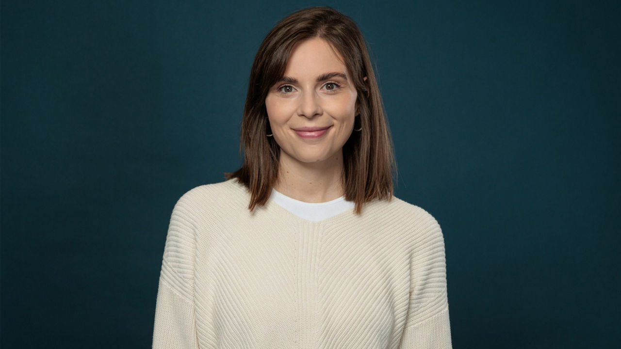 A portrait of a person (Sarah Hinderer) against a dark blue-green background. The person is wearing a cream-colored knit sweater over a white top and silver hoop earrings. Shoulder-length brown hair frames the face. The person is looking directly at the camera. The image is styled as a professional business portrait with even lighting and a simple background.