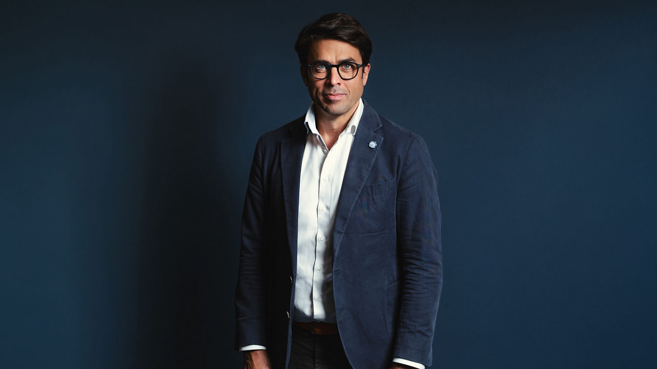 Christian Schmitz: A man with short dark hair and glasses is standing in front of a dark blue background. He is wearing a white shirt and a navy blazer with a small pin on the lapel, and he looks directly at the camera with a neutral expression.