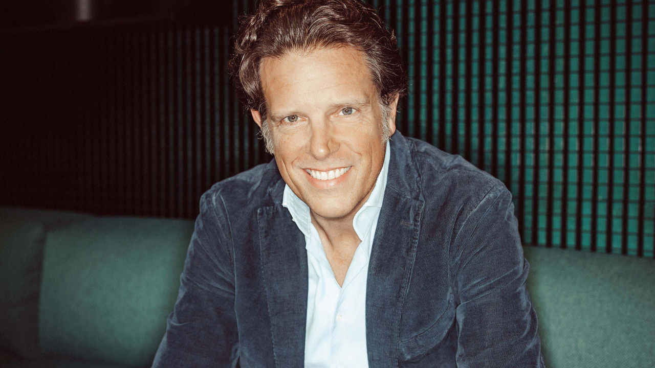 Florian Haller is smiling at the camera while sitting on a green sofa in a modern, dimly lit room. He is wearing a light blue shirt and a dark blue corduroy jacket. The background features vertical green-black acoustic panels.
