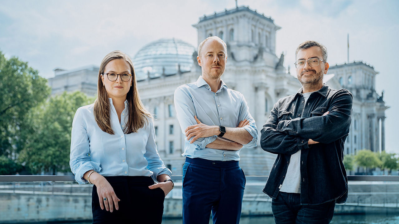 Das Bild zeigt drei Personen, Anna Brockmann, Johannes Wendlinger und Benedikt Göttert, – zwei Männer und eine Frau – in Businesskleidung, die vor dem Reichstagsgebäude in Berlin stehen und selbstbewusst in die Kamera blicken.