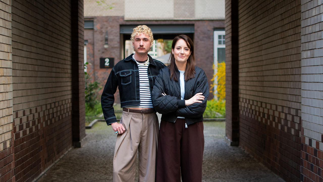 Zwei Personen stehen zwischen Backsteinwänden in einem Innenhof und blicken in die Kamera.
