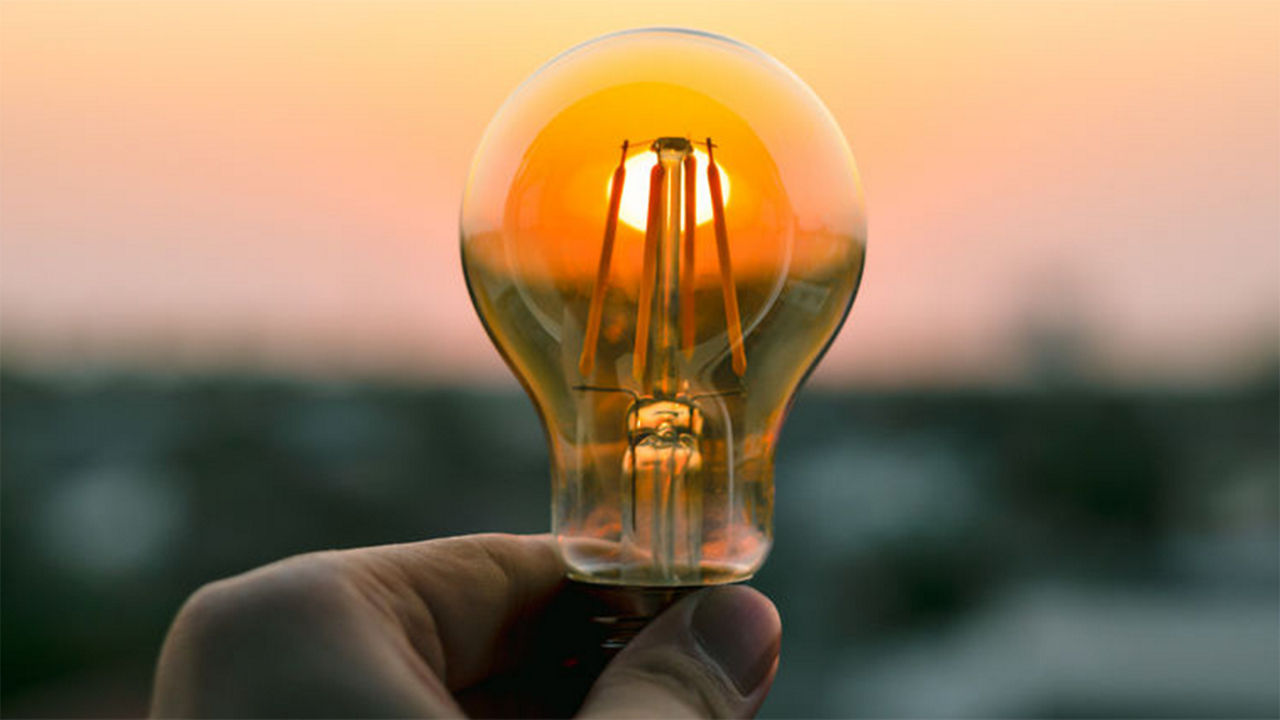 Hand hält eine Glühbirne, in der sich die untergehende Sonne spiegelt.