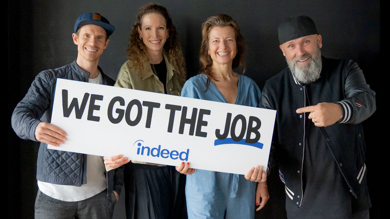 Andy Wyeth, Liselotte Schwenkert, Stefanie Kuhnhen und Niko Backspin halten ein Schild mit der Aufschrift „WE GOT THE JOB – indeed“ und lächeln in die Kamera.