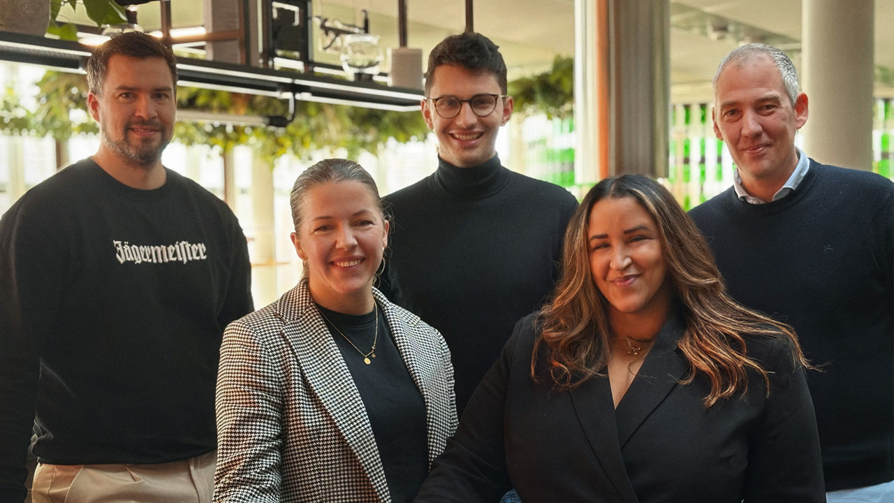 Fünf Personen stehen nebeneinander in einem Innenraum und blicken in die Kamera.