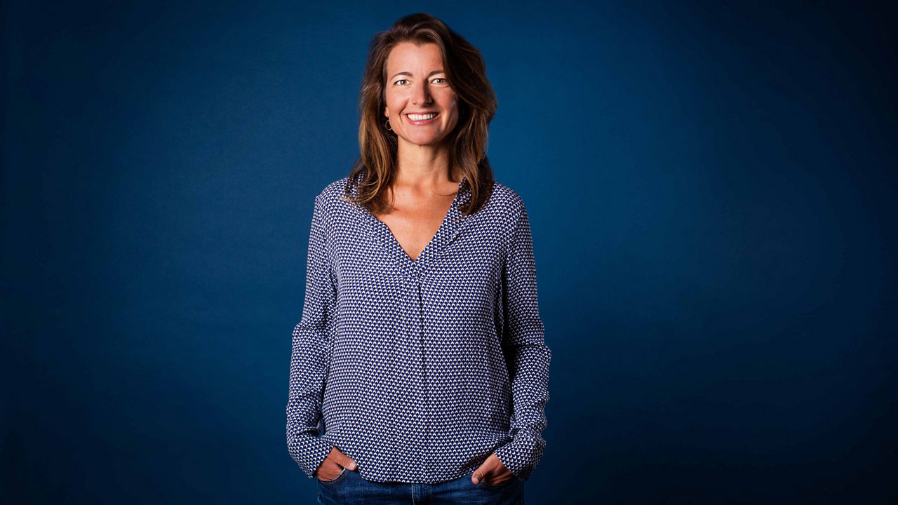Porträtfoto von Stefanie Kuhnhen vor blauem Hintergrund. Sie trägt eine gemusterte Bluse, lächelt in die Kamera und hat die Hände in den Hosentaschen.