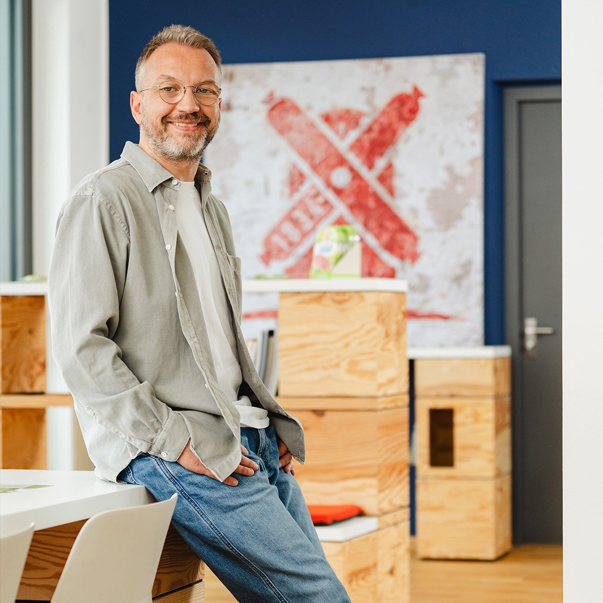 Steffen Zeller: Ein Mann mittleren Alters mit Brille, grauem Vollbart und kurzen Haaren steht locker an einen Tisch gelehnt in einem modernen Büro mit Holzboden. Er trägt ein hellgraues Hemd über einem weißen T-Shirt, eine locker sitzende Jeans und graue New-Balance-Sneaker. Im Hintergrund ist ein modernes Kunstwerk mit roten Elementen zu sehen, das auf eine Wurstmarke hinweist. Der Mann schaut freundlich und direkt in die Kamera, mit leichtem Lächeln. Die Szenerie wirkt entspannt und einladend.