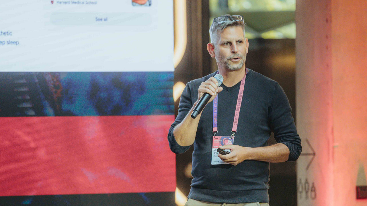 The image shows a middle-aged man with short grey hair speaking on a stage. He is wearing a dark long-sleeve shirt, has glasses resting on his head, and wears a pink lanyard with a badge labeled "Innovation." He is holding a microphone in his right hand and a smartphone in his left. In the background, a large digital screen with blurred text and visuals is visible, along with a lit wall featuring a red color gradient. The setting appears to be a professional event or conference. The speaker appears focused and engaged. This alt text is written for accessibility according to WCAG standards and optimized for SEO terms such as innovation keynote speaker, conference presentation, and tech events.