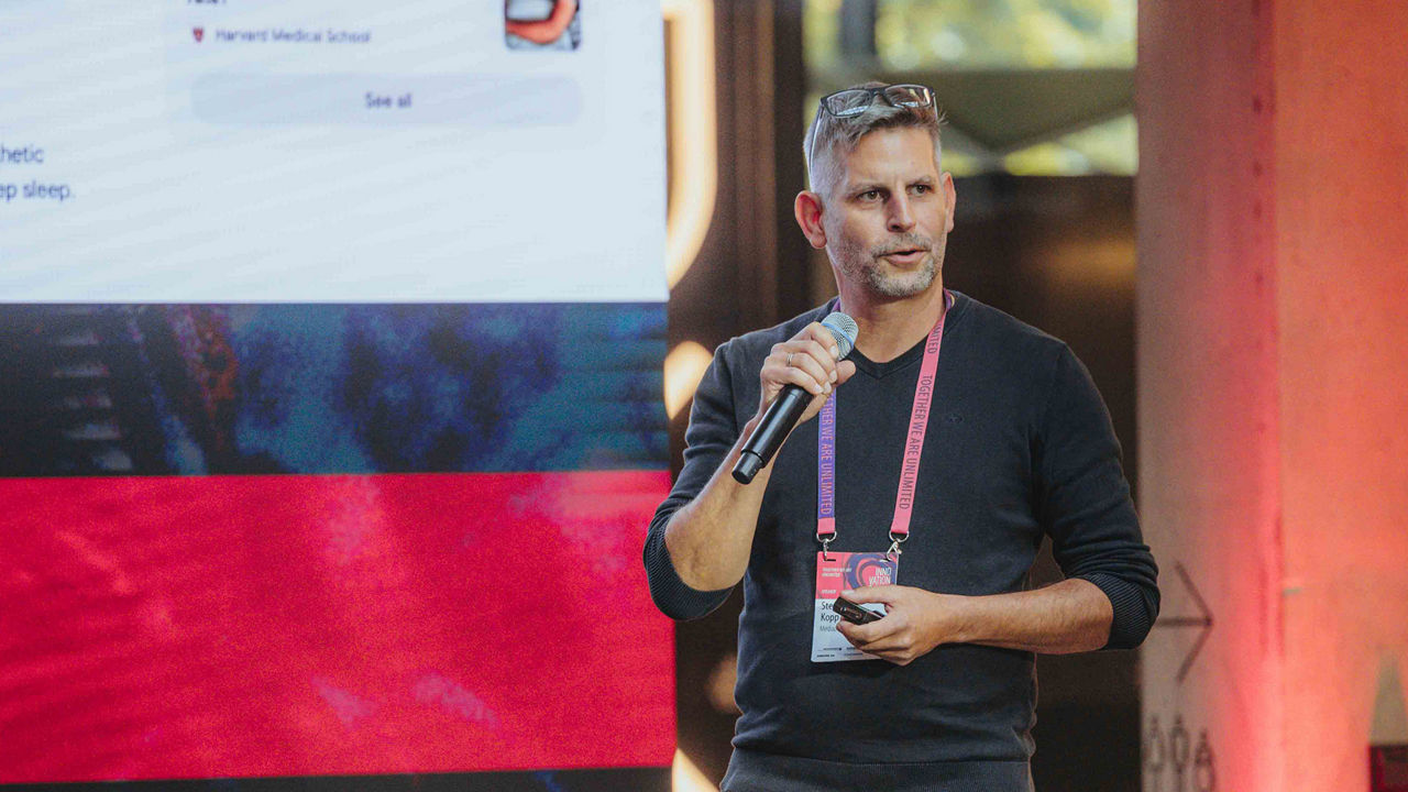 A person (Stephan Kopp) holding a microphone is giving a presentation on stage in front of a large screen display at an event.