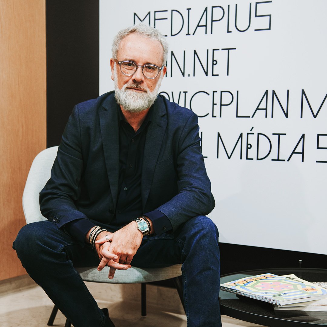 Mann mit grauem Bart und Brille sitzt in einem modernen Bürobereich vor einer Wand mit Firmenlogos. Er trägt einen dunklen Anzug und blickt ruhig in die Kamera.