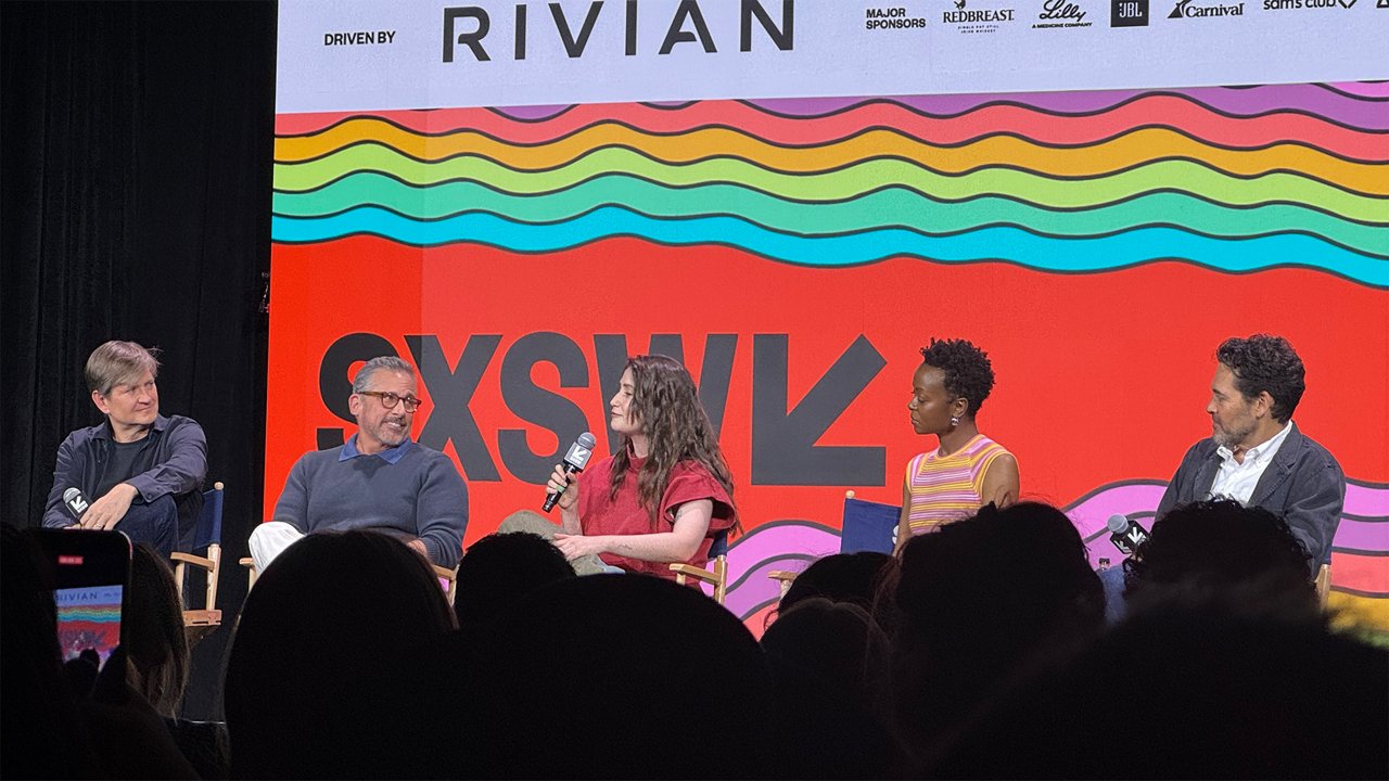 Panel discussion with five people on a SXSW stage in front of a large colorful screen displaying the SXSW logo. One person speaks into a microphone while the others listen, with the audience visible in the foreground.